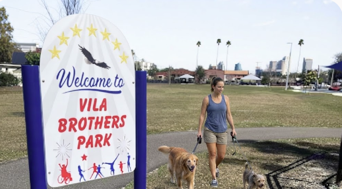 A person walking two leashed dogs beside the Welcome to Vila Brothers Park sign.